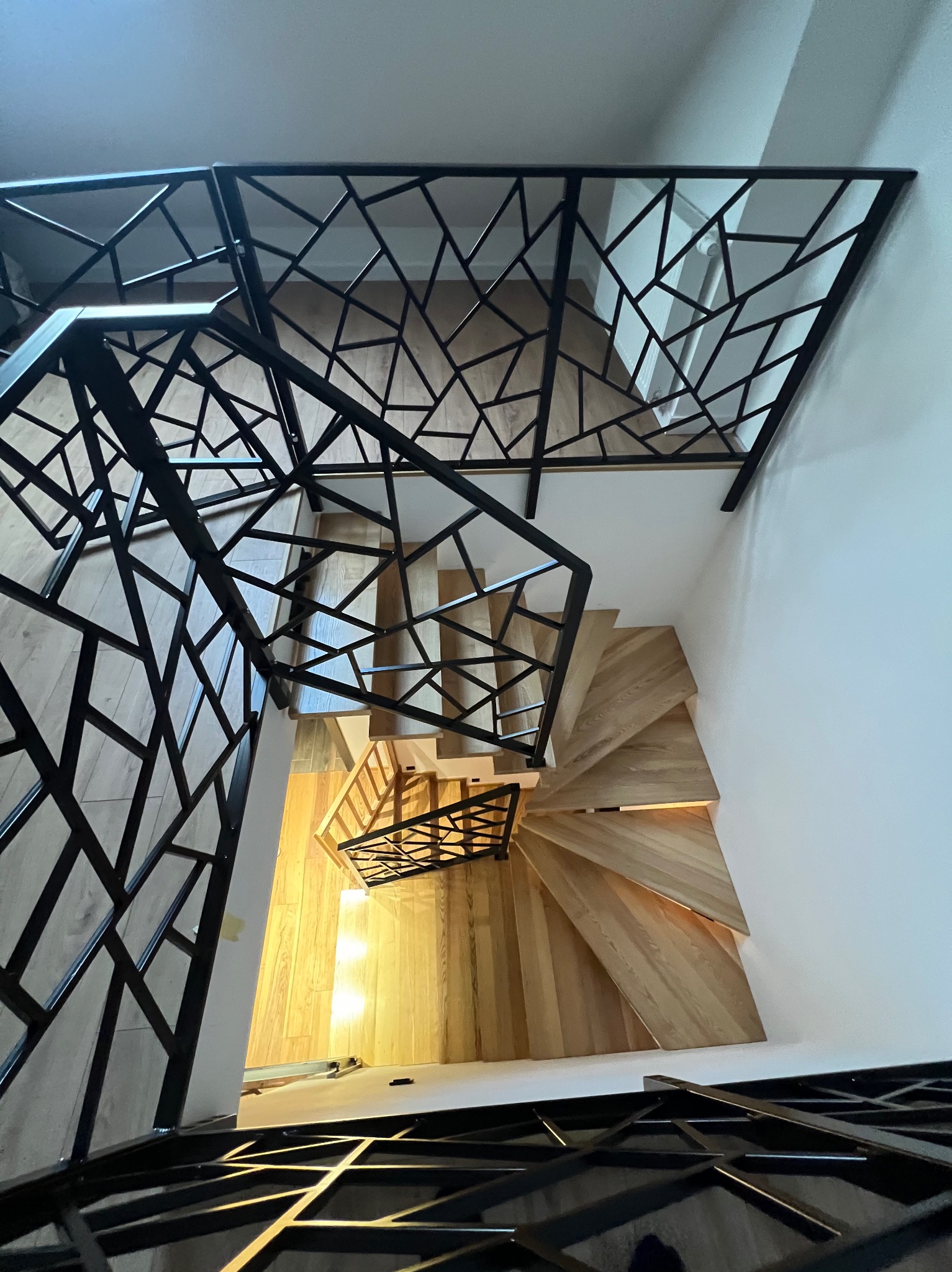 Spiralne schody z drewnianymi stopniami i nowoczesną, geometryczną, czarną konstrukcją. Widok z góry, podkreślający unikalny design i jasne oświetlenie.