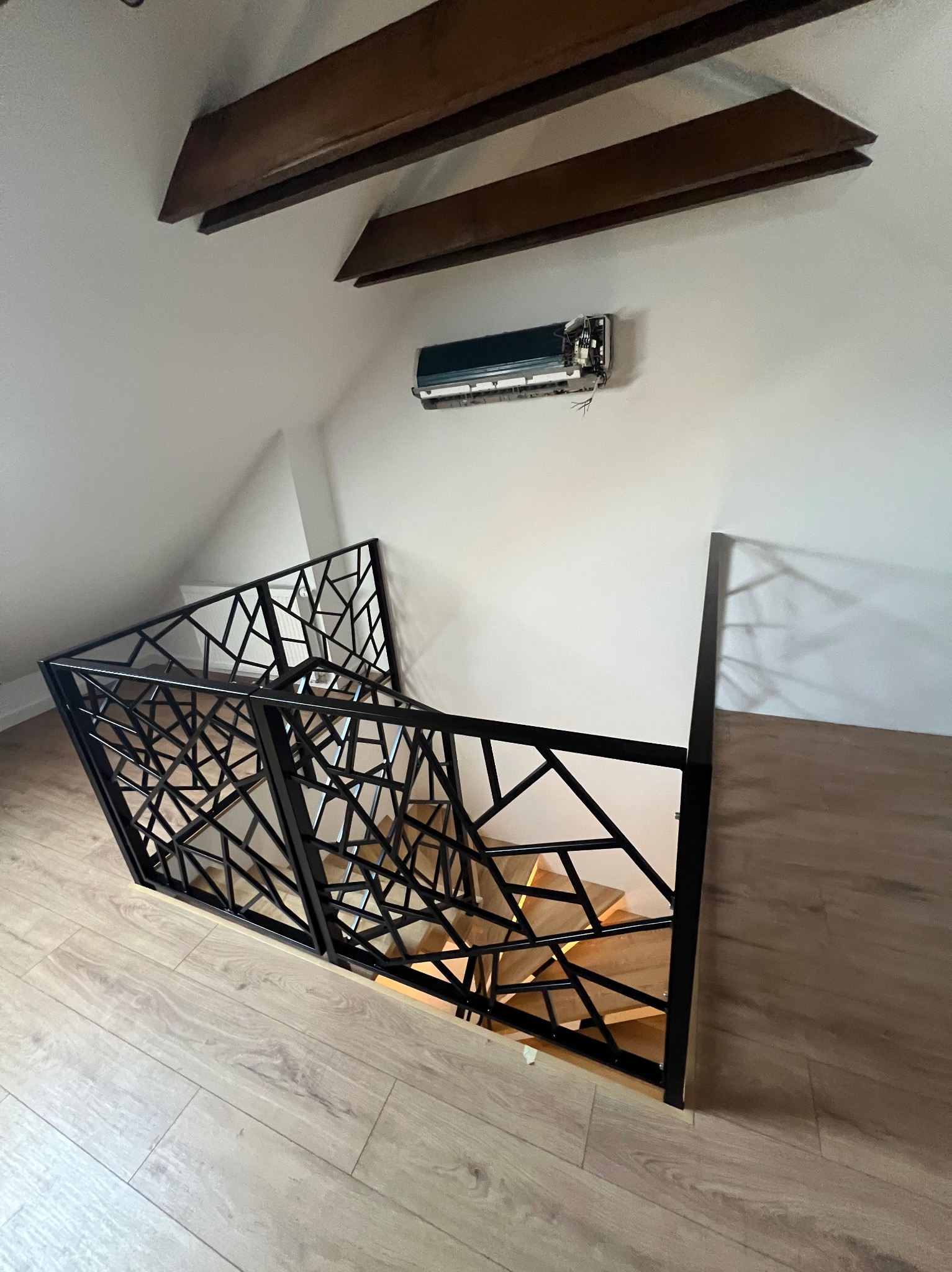 Nowoczesna, czarna barierka schodowa z geometrycznym wzorem, zamontowana w jasnym wnętrzu z drewnianymi schodami i podłogą. Widok z góry na dół.