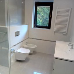 Remont - Nowoczesna łazienka w odcieniach bieli i szarości z toaletą, bidetem, umywalką z szafką, grzejnikiem drabinkowym i oknem z widokiem na zieleń.