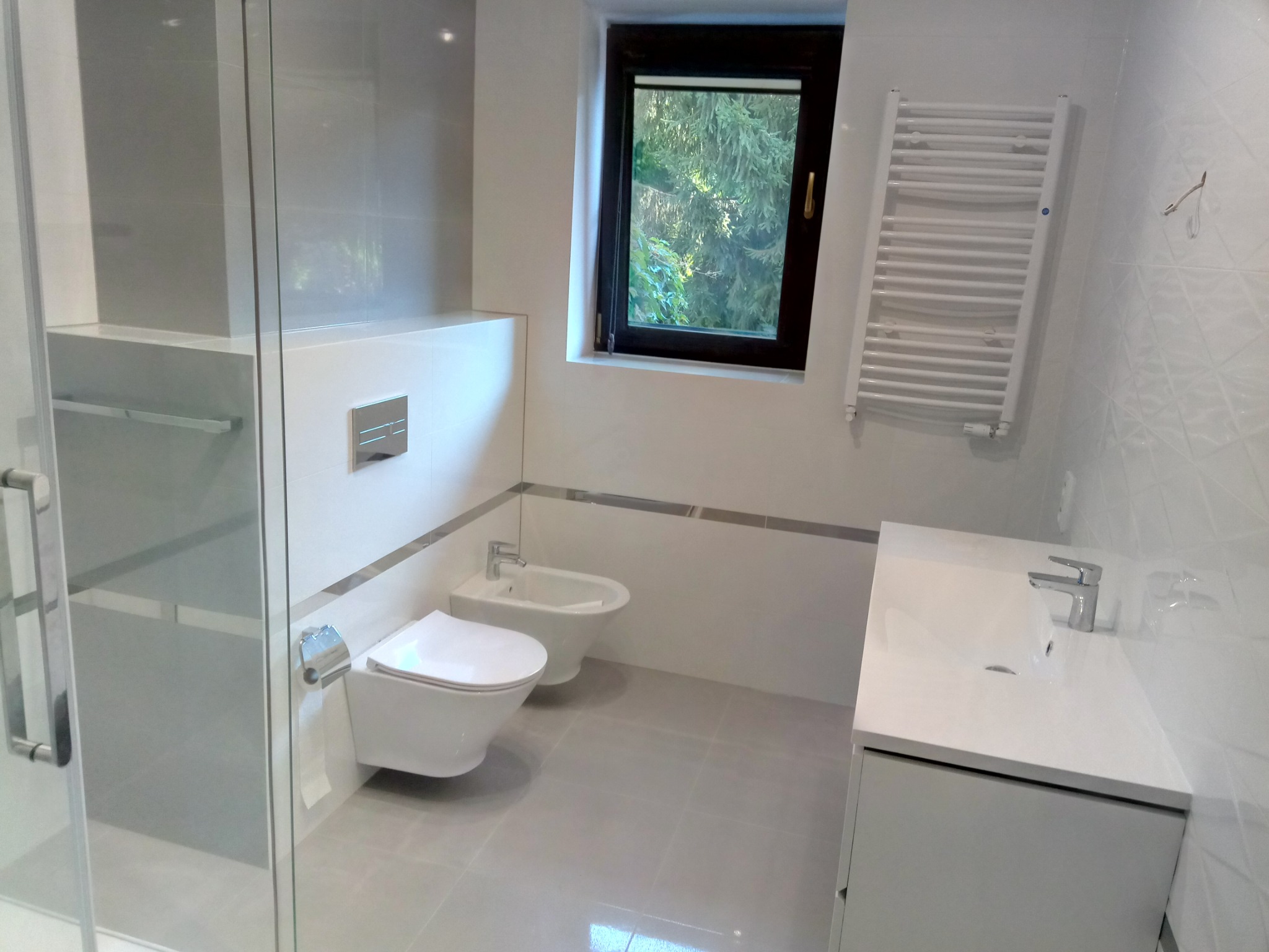 Nowoczesna łazienka w odcieniach bieli i szarości z toaletą, bidetem, umywalką z szafką, grzejnikiem drabinkowym i oknem z widokiem na zieleń.