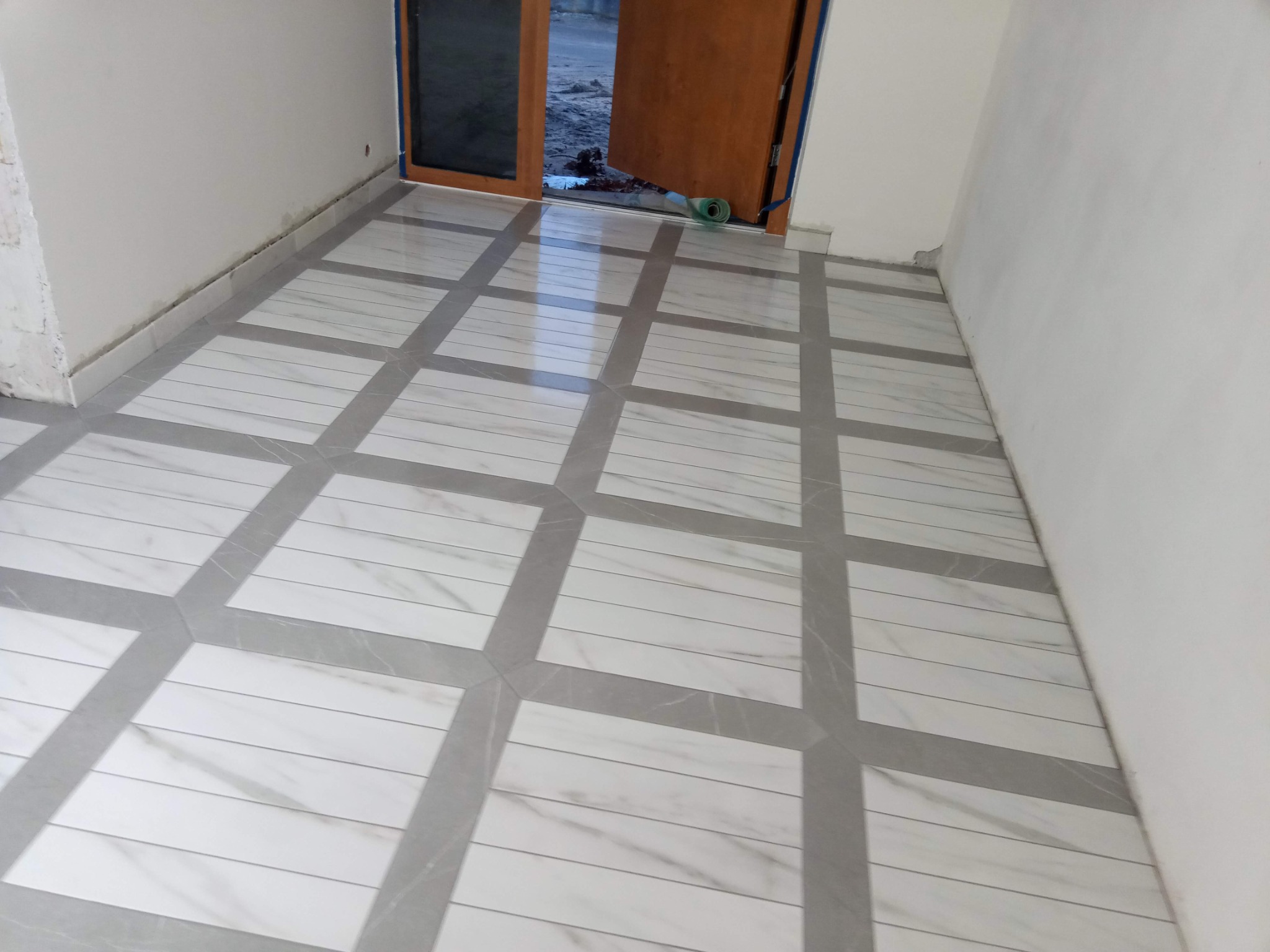Ułożona podłoga z płytek imitujących marmur w geometryczny wzór z szarymi fugami w nowym pomieszczeniu z otwartymi drewnianymi drzwiami.