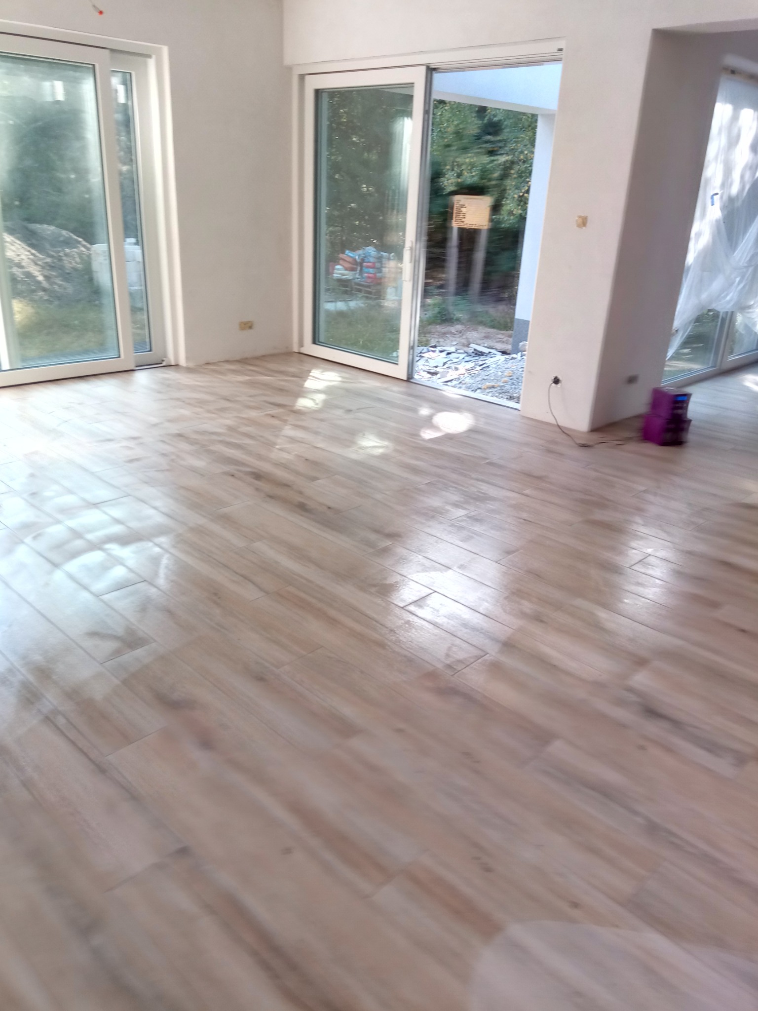 Jasne, drewnopodobne panele ułożone w przestronnym wnętrzu z dużymi oknami przesuwnymi i widokiem na zieleń na zewnątrz. Pod ścianą widoczny fioletowy sprzęt budowlany.