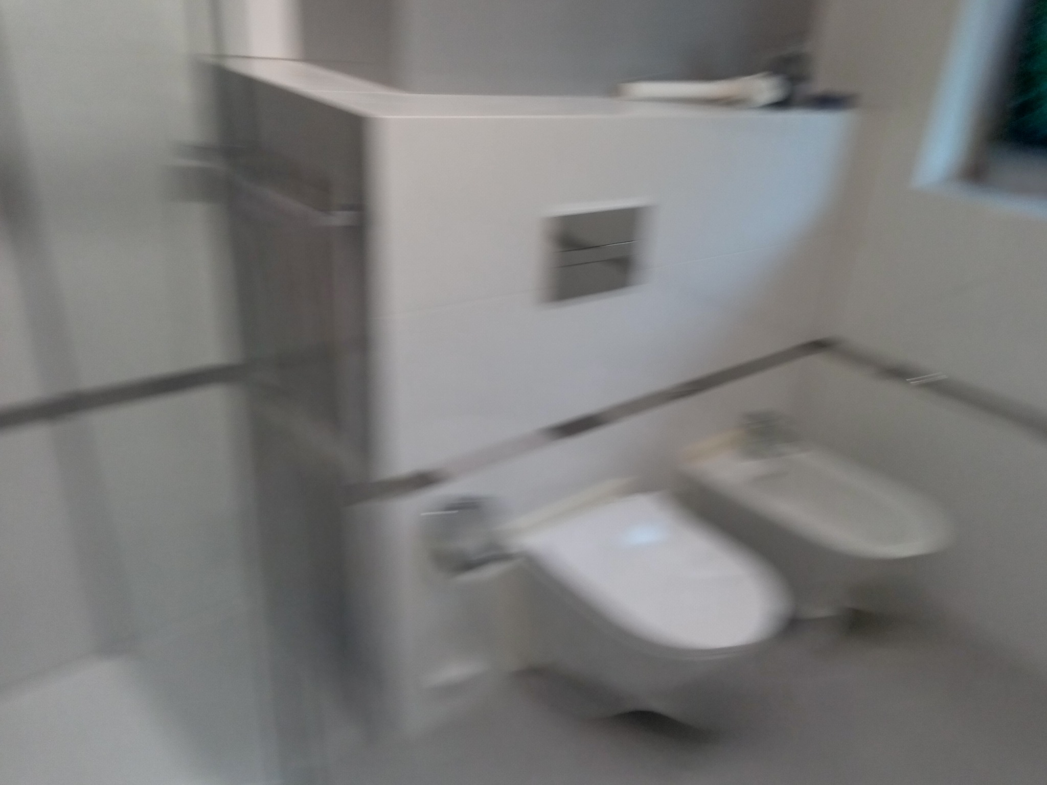 Nowoczesna, minimalistyczna łazienka z białymi płytkami, podwieszaną miską WC i bidetem; widok zza szklanej kabiny prysznicowej, z rozmytym efektem ruchu.