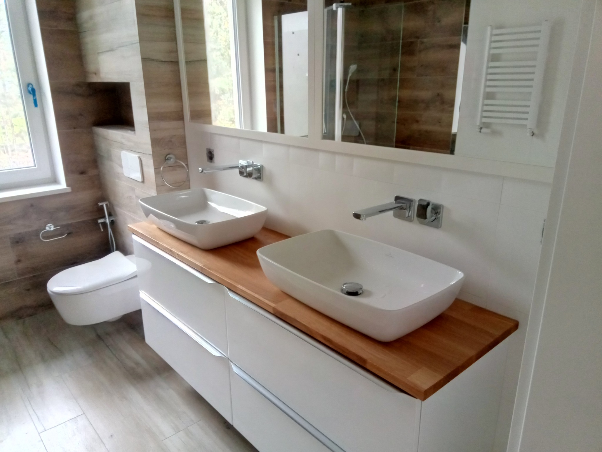 Nowoczesna łazienka z dwoma białymi umywalkami nablatowymi na drewnianym blacie, białe szafki pod umywalkami, toaleta podwieszana, grzejnik drabinkowy i lustro na ścianie wyłożonej płytkami...