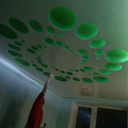 PG-AGRA SPÓŁKA Z OGRANICZONĄ ODPOWIEDZIALNOŚCIĄ - Biały sufit podwieszany z zielonymi okrągłymi wzorami w kształcie spirali, oświetlony od boku zielonym światłem, z widoczną postacią sięgającą do sufitu.