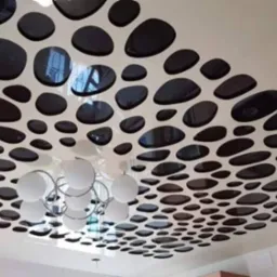 Nowoczesny sufit podwieszany z dekoracyjnymi, owalnymi otworami w kolorze czarnym, przez które widać odbicia, z widocznym fragmentem żyrandola z białymi kulami.