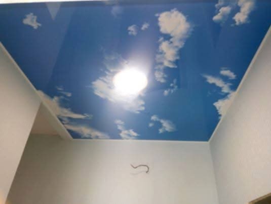 Podwieszany sufit z nadrukiem błękitnego nieba i obłoków w jasnym pomieszczeniu, widoczne białe ściany i element instalacji na ścianie.
