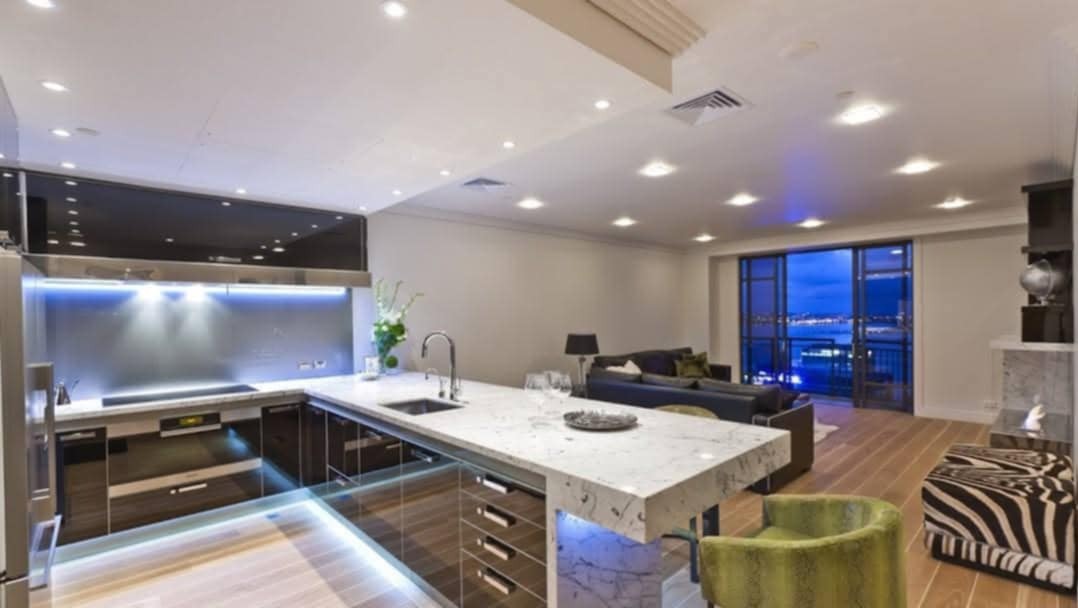 Nowoczesna kuchnia z wyspą o marmurowym blacie, podświetleniem LED i widokiem na miasto za oknem.