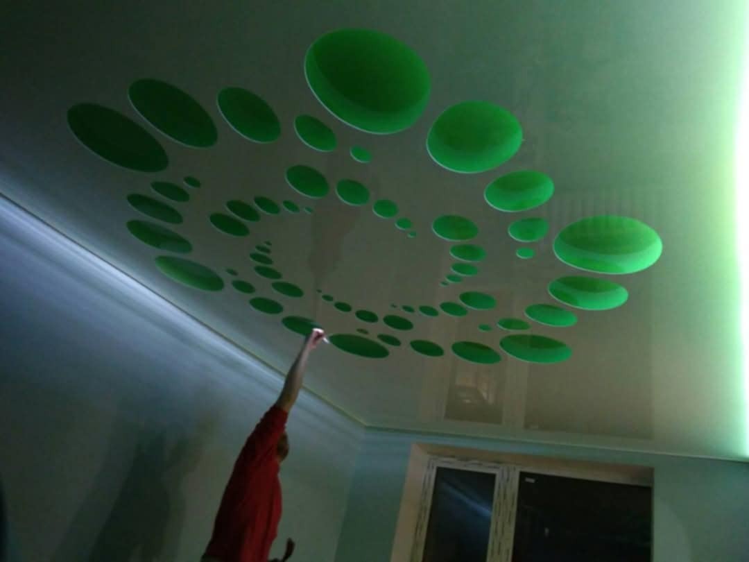 Biały sufit podwieszany z zielonymi okrągłymi wzorami w kształcie spirali, oświetlony od boku zielonym światłem, z widoczną postacią sięgającą do sufitu.