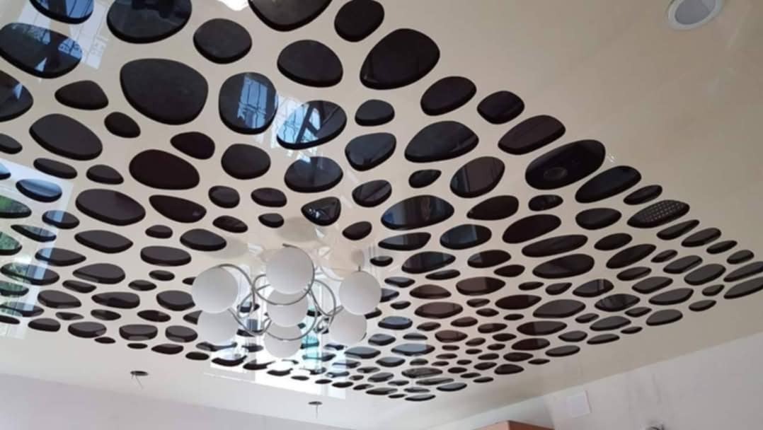 Nowoczesny sufit podwieszany z dekoracyjnymi, owalnymi otworami w kolorze czarnym, przez które widać odbicia, z widocznym fragmentem żyrandola z białymi kulami.