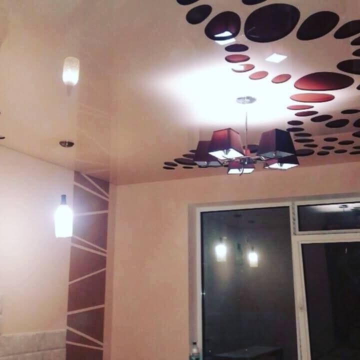 Pokój z kremowymi ścianami, sufitem z dekoracyjnymi okręgami i nowoczesnym oświetleniem: wisząca lampa z abażurami i podświetlane butelki na ścianie.