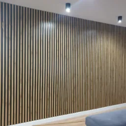 Drewniana ściana z pionowych listew w nowoczesnym wnętrzu, z widocznym fragmentem szarej sofy i dwoma czarnymi lampami sufitowymi.