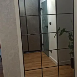 Duże lustro w czarnej ramie z podziałami, odbijające fragment przedpokoju z drewnianą podłogą, lampą sufitową i rośliną w doniczce.