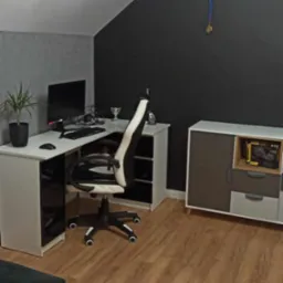 Pokój na poddaszu z drewnianą podłogą, biurkiem narożnym z komputerem, krzesłem gamingowym biało-czarnym, komodą i ścianami w kolorze szarym i grafitowym, udekorowany medalem na ścianie.