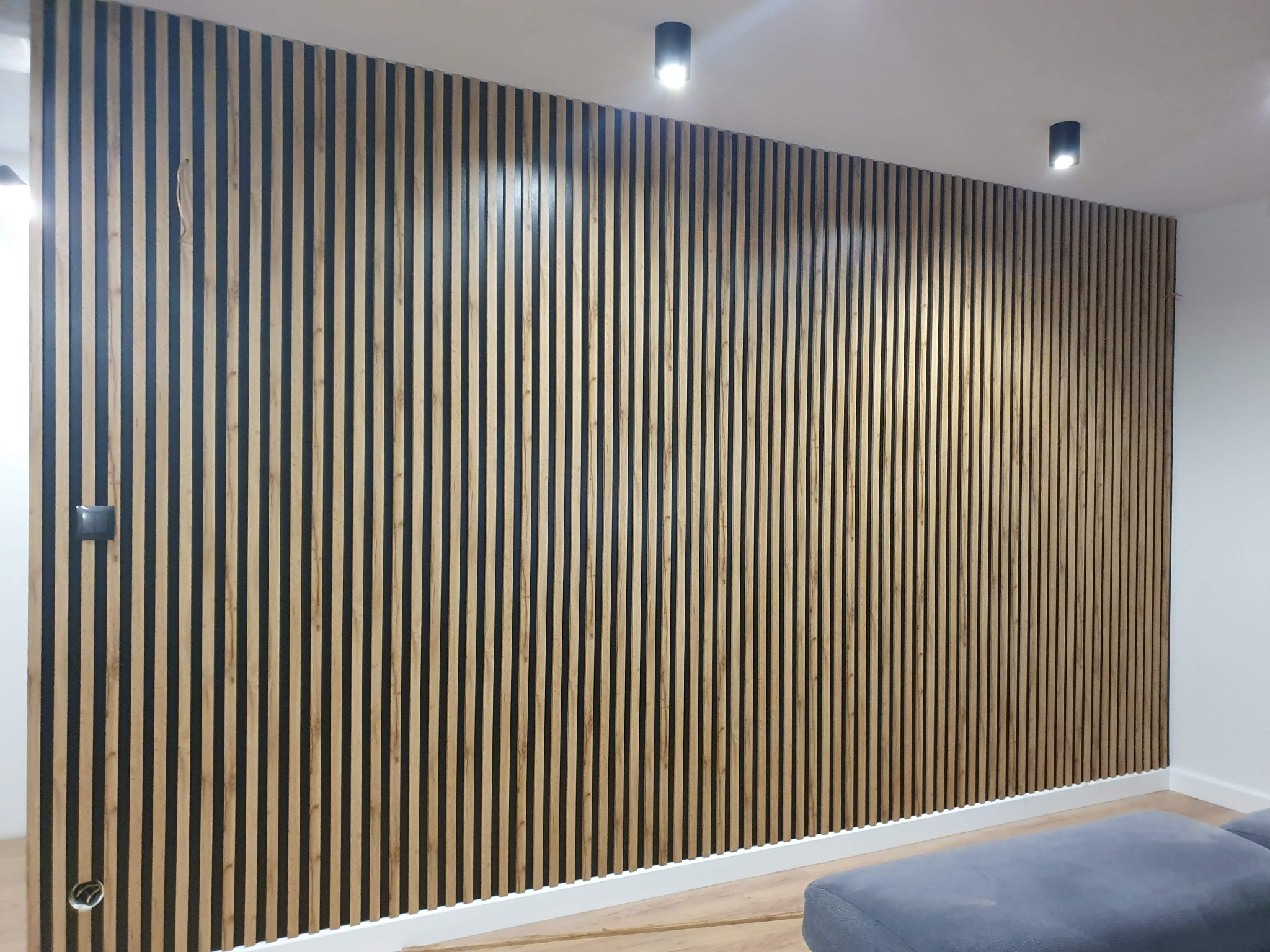 Drewniana ściana z pionowych listew w nowoczesnym wnętrzu, z widocznym fragmentem szarej sofy i dwoma czarnymi lampami sufitowymi.