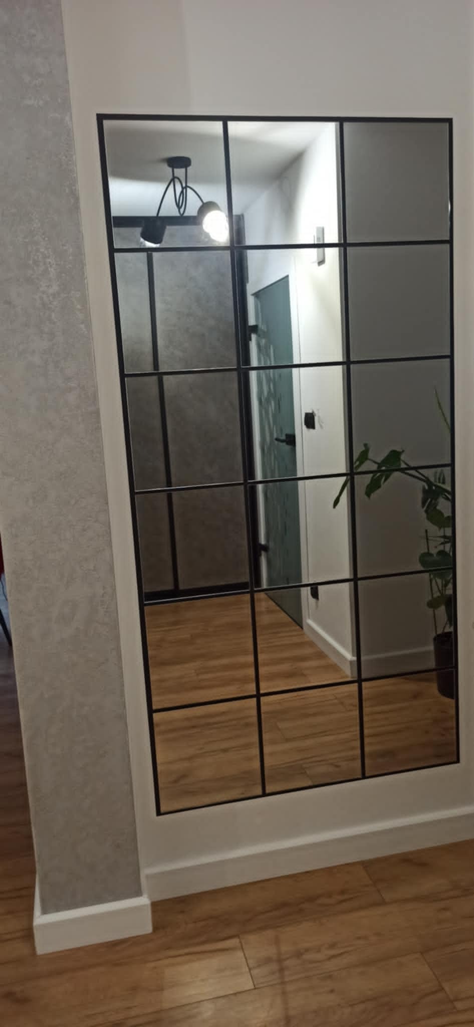 Duże lustro w czarnej ramie z podziałami, odbijające fragment przedpokoju z drewnianą podłogą, lampą sufitową i rośliną w doniczce.