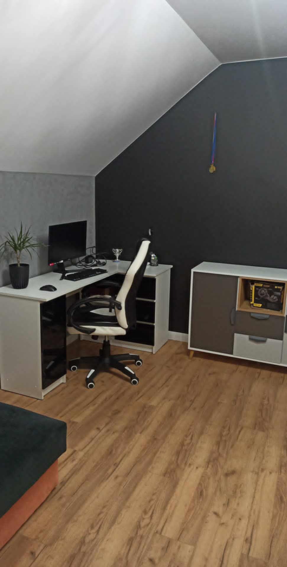 Pokój na poddaszu z drewnianą podłogą, biurkiem narożnym z komputerem, krzesłem gamingowym biało-czarnym, komodą i ścianami w kolorze szarym i grafitowym, udekorowany medalem na ścianie.