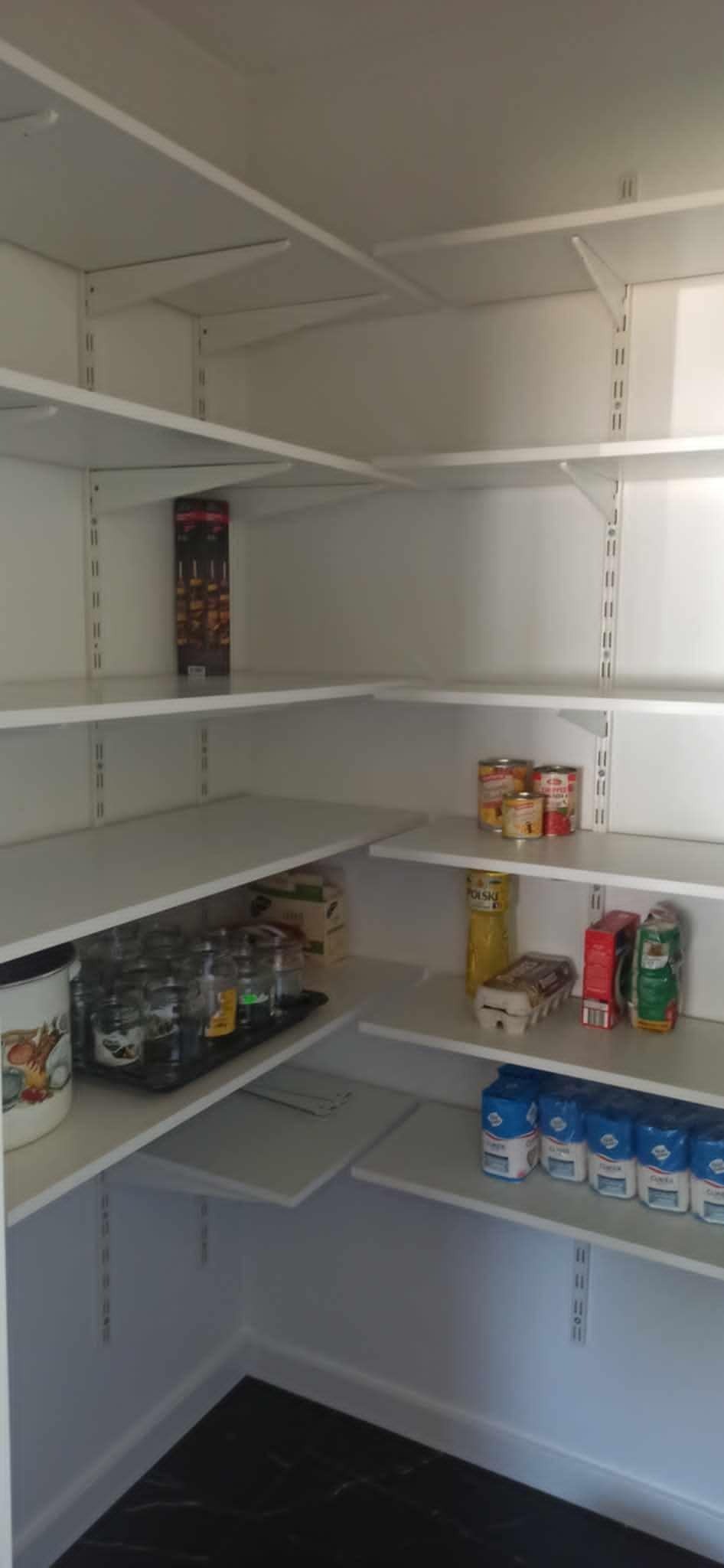 Biała, pusta spiżarnia z regulowanymi półkami w Łańcucie, częściowo wypełniona produktami spożywczymi, takimi jak mąka, puszki i jajka.