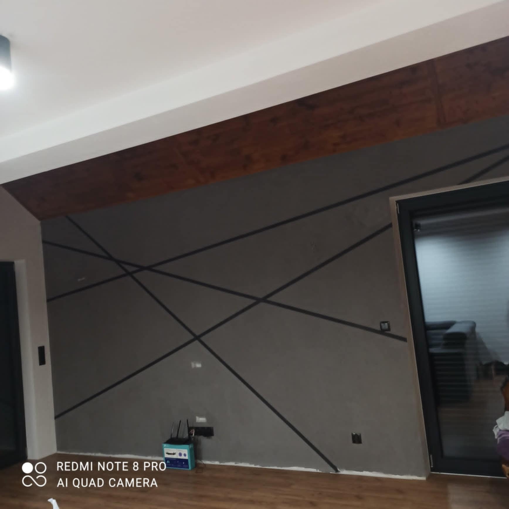 Pomieszczenie z szarą ścianą ozdobioną czarnymi, ukośnymi liniami, drewnianą belką pod sufitem i podłogą z paneli. Widoczny router i gniazdka elektryczne.