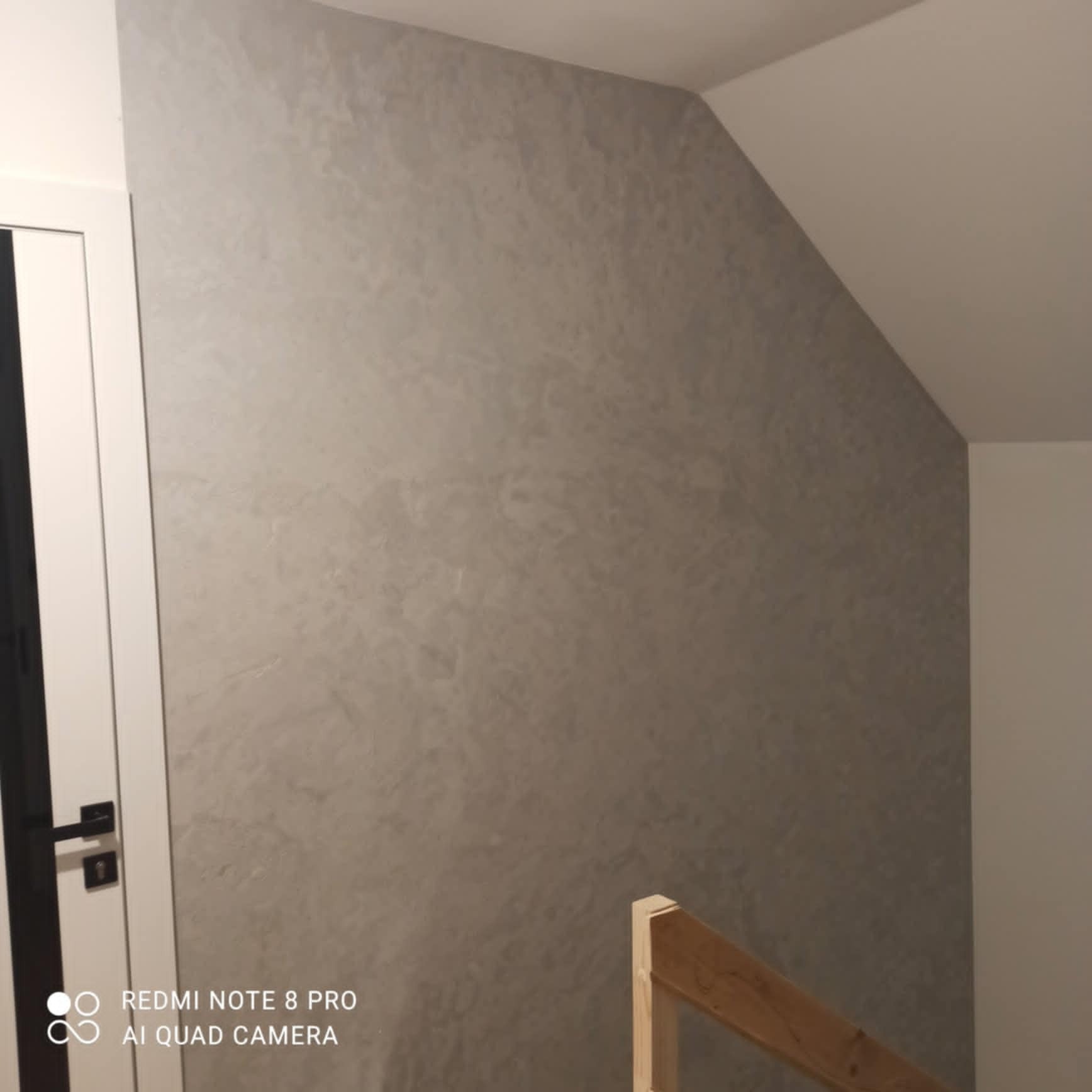 Ściana w odcieniach szarości z efektem dekoracyjnym, widoczna część białej stolarki drzwiowej i drewniana balustrada schodów w tle.