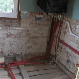 Surowa ściana z cegły i nowa instalacja hydrauliczna z czerwonymi rurami, widoczny fragment okna i podłoga w trakcie remontu.