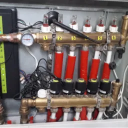 Rozdzielacz ogrzewania podłogowego w szafce, z numerowanymi sekcjami (1-6), czerwonymi rurami i czarnymi głowicami termostatycznymi. Widoczny manometr i przewody.