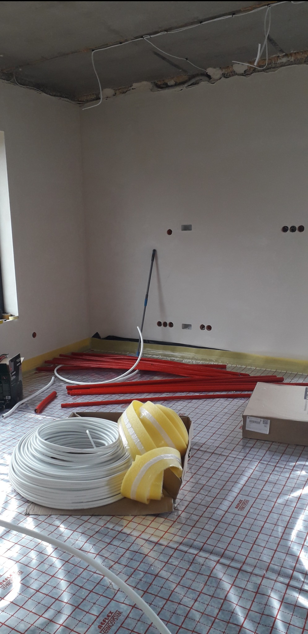 Przygotowanie do montażu ogrzewania podłogowego: rozłożona mata grzewcza z siatką, zwój białego przewodu, czerwone rurki i żółta taśma izolacyjna w surowym wnętrzu z widocznymi przewodami w suficie.