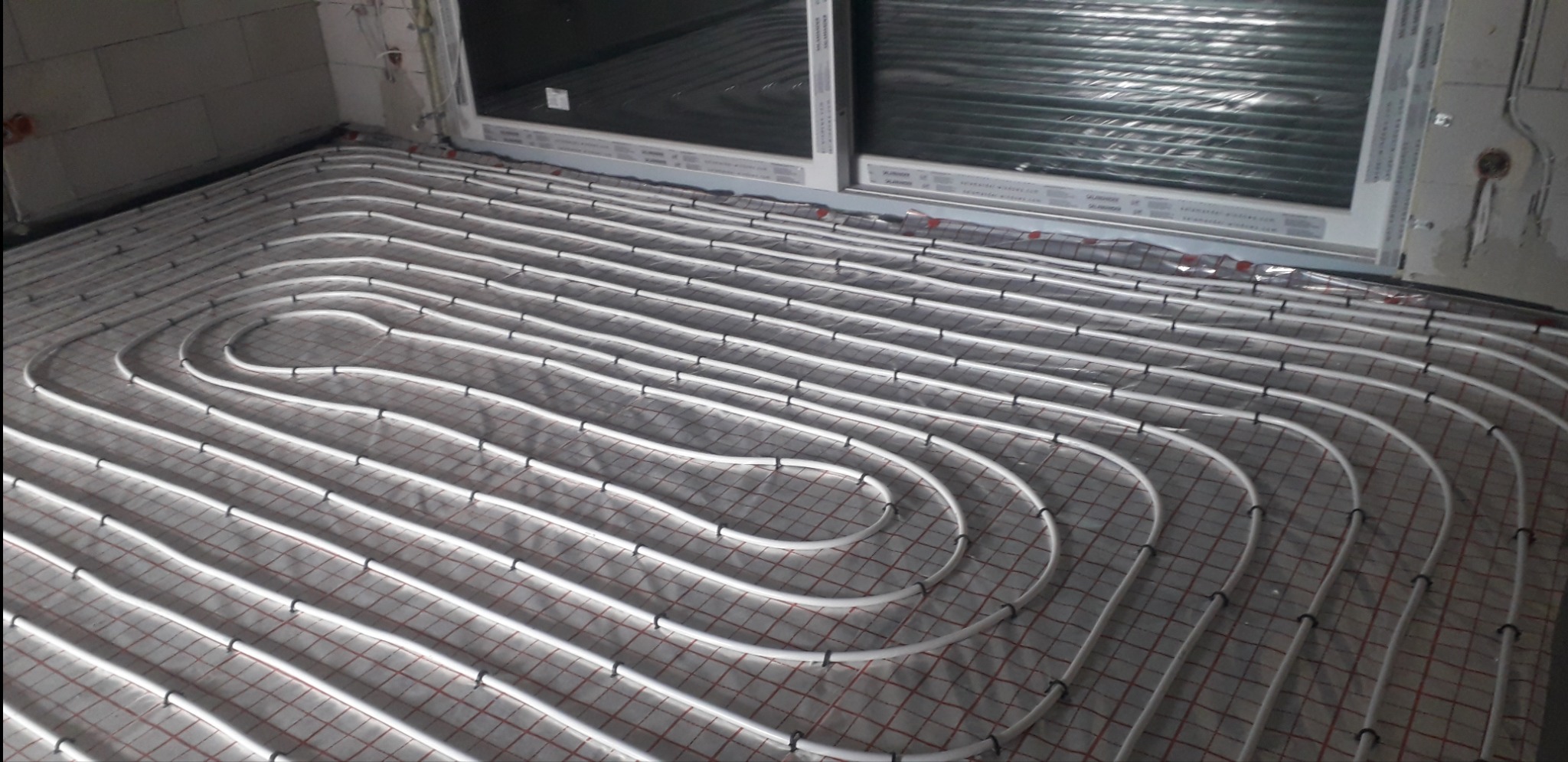 Białe rurki ogrzewania podłogowego ułożone w pętle na folii z czerwoną siatką, widoczne okno i fragment ściany z instalacjami elektrycznymi.