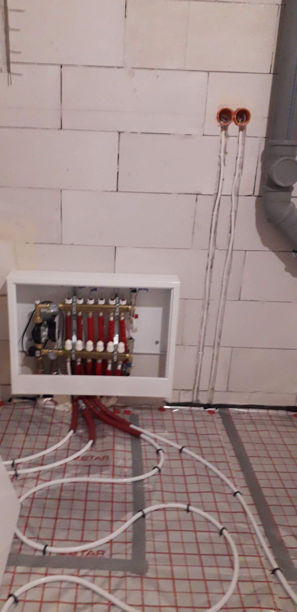 Rozdzielacz ogrzewania podłogowego w białej szafce, rury grzewcze ułożone na folii z nadrukowaną siatką, ściana z białego betonu komórkowego, widoczne kable elektryczne i fragment rury kanalizacyjnej.