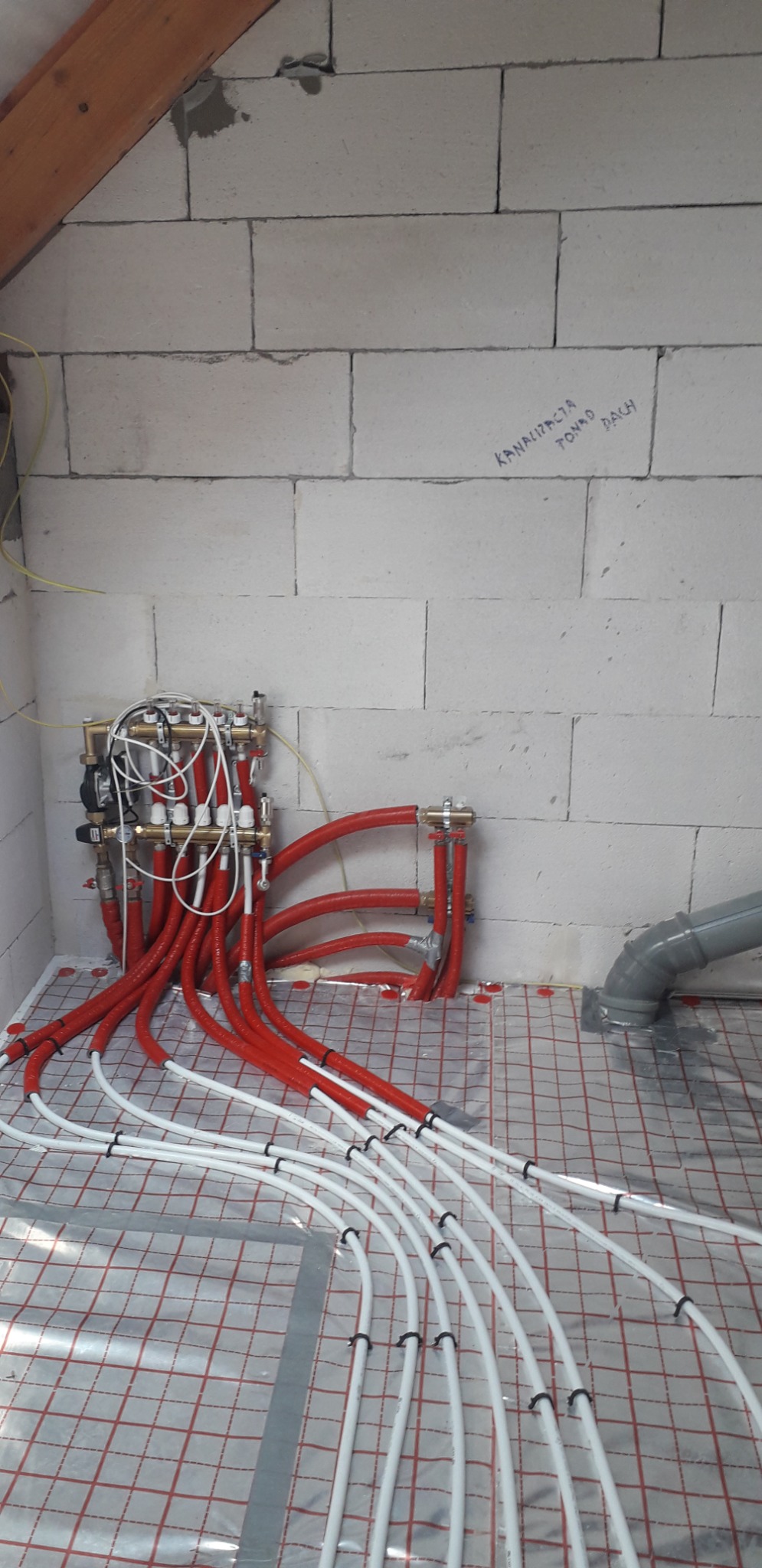 Rozdzielacz ogrzewania podłogowego z czerwonymi i białymi rurami na folii grzewczej, widoczna ściana z bloczków z napisem 'Kanalizacja Tomko Dach' i drewniana belka stropowa.
