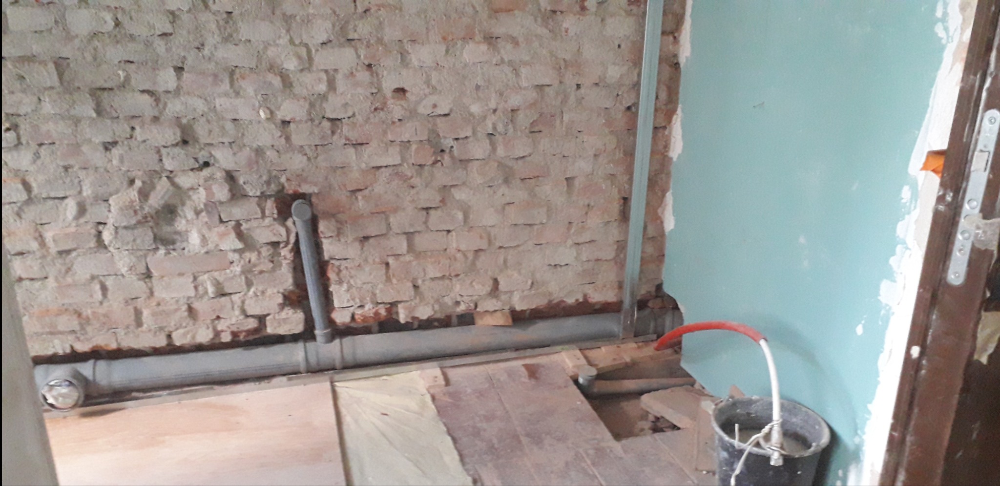 Wnętrze w trakcie remontu: odsłonięta ceglana ściana, szare rury instalacji hydraulicznej biegnące wzdłuż ściany, fragment podłogi z płyt, niebieska ściana z widocznymi śladami prac wykończeniowych...