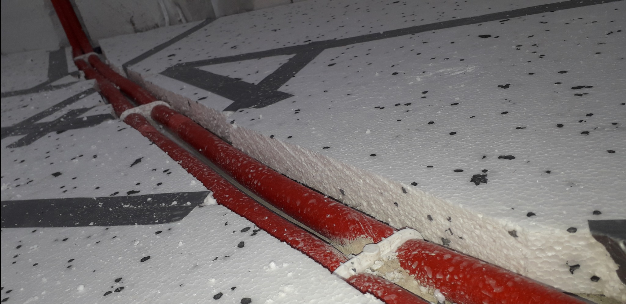 Czerwone rury grzewcze ułożone w wycięciu w styropianie pod ogrzewanie podłogowe, widoczne czarne plamki na izolacji i szare oznaczenia na podłożu.