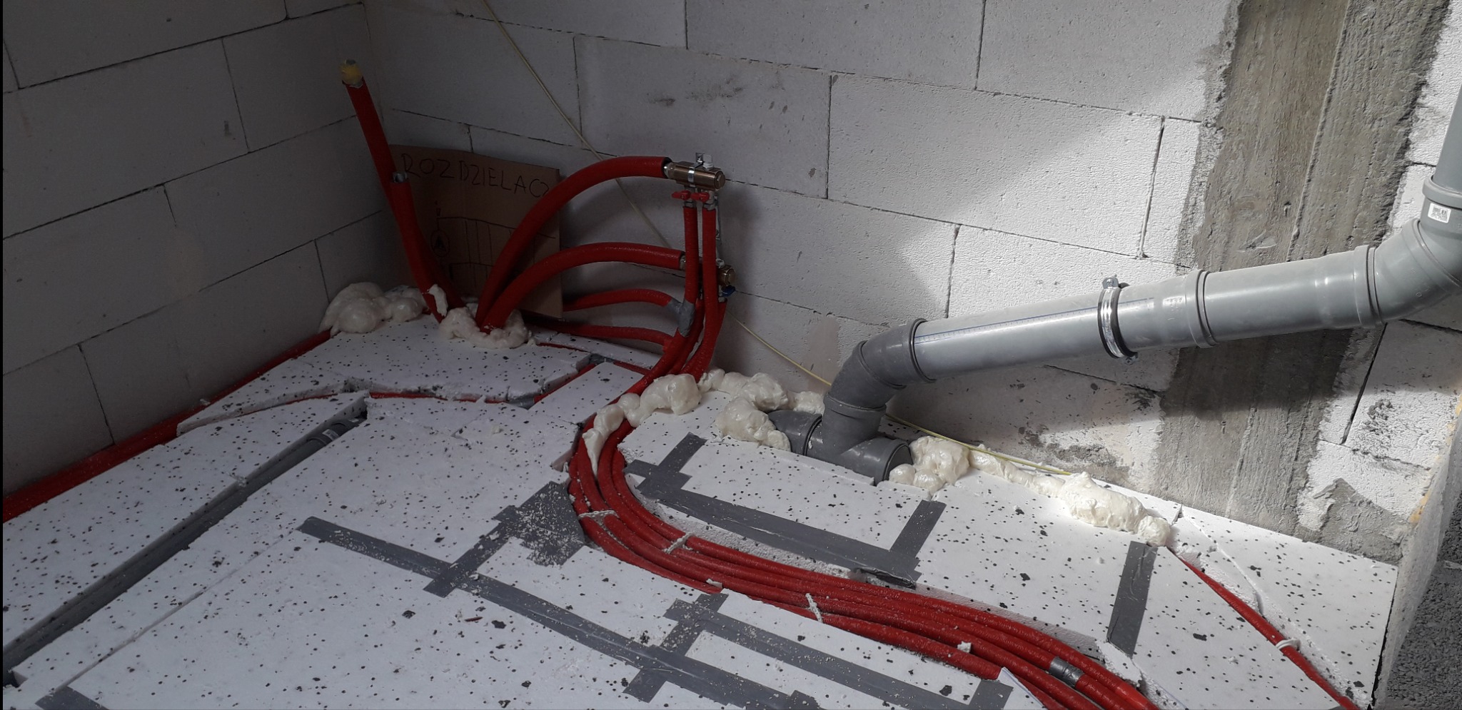Czerwone rury ogrzewania podłogowego zalane pianką montażową na izolacji podłogowej, szare rury kanalizacyjne w rogu pomieszczenia z surowymi ścianami z betonu komórkowego.