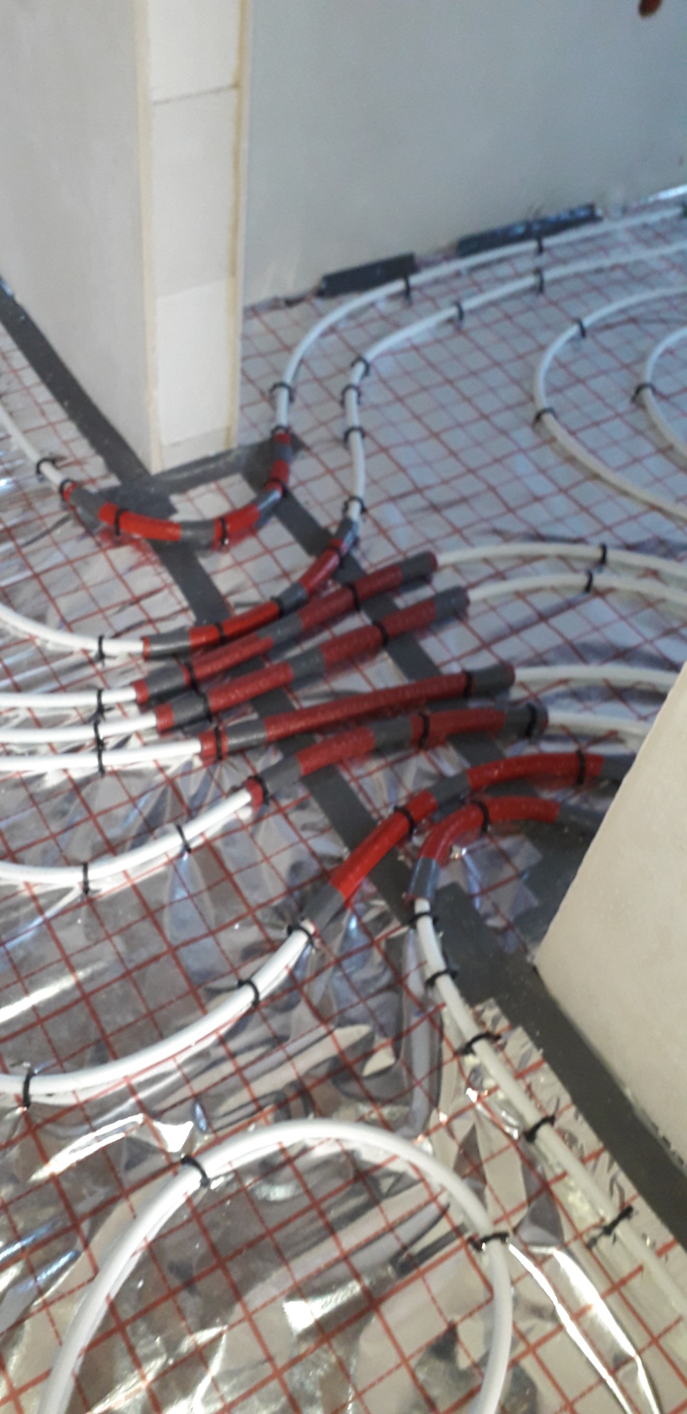 Białe rury ogrzewania podłogowego na folii izolacyjnej z czerwono-szarymi złączkami, przymocowane czarnymi opaskami zaciskowymi, widok z góry podczas instalacji.