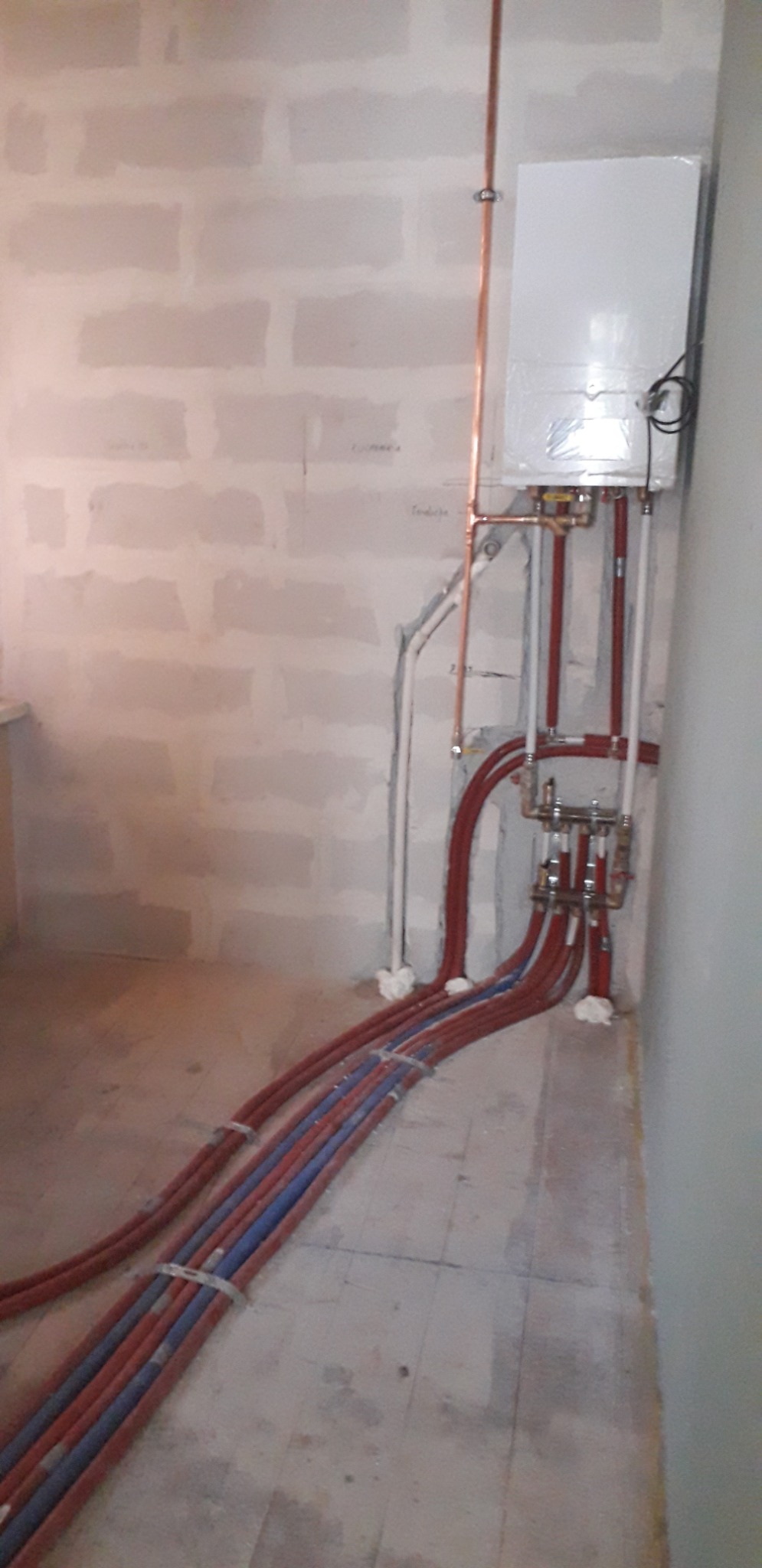 Nowy piec gazowy w folii ochronnej, z podłączonymi rurami miedzianymi i instalacją ogrzewania podłogowego, widok na surową ścianę z niedokończonymi tynkami.