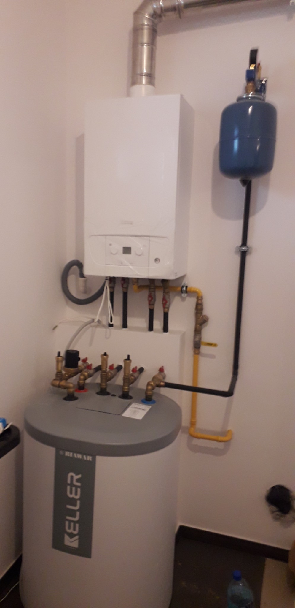 Biały piec gazowy kondensacyjny z szarym zasobnikiem wody marki Keller i niebieskim naczyniem przeponowym, rury w kolorze żółtym i czarnym, zamontowane na ścianie w pomieszczeniu.