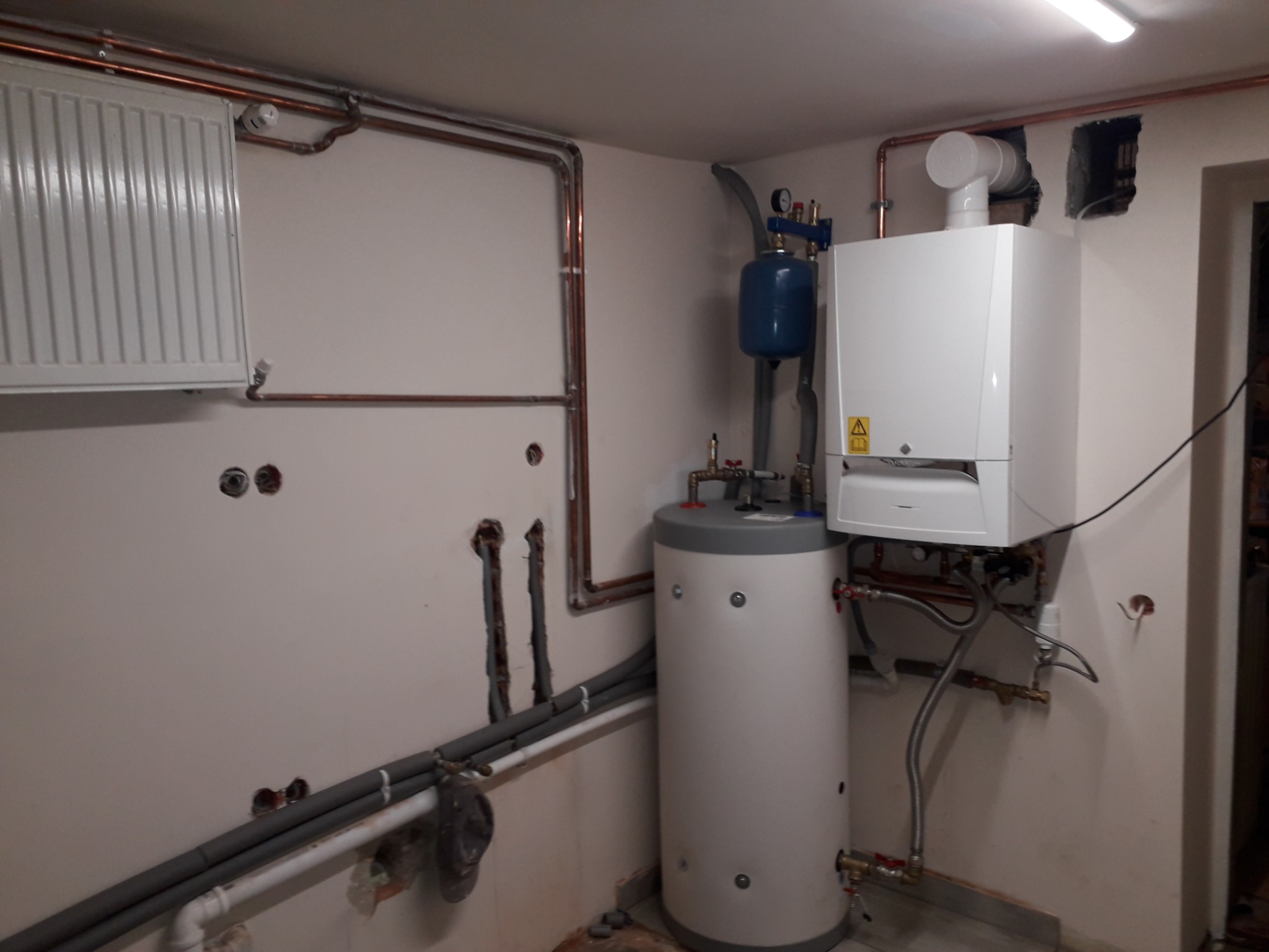 Nowoczesny, biały piec gazowy zamontowany na ścianie, obok pionowy zasobnik na wodę, widoczne miedziane rury instalacji grzewczej i szare rury doprowadzające wodę, surowy stan wykończenia ścian.