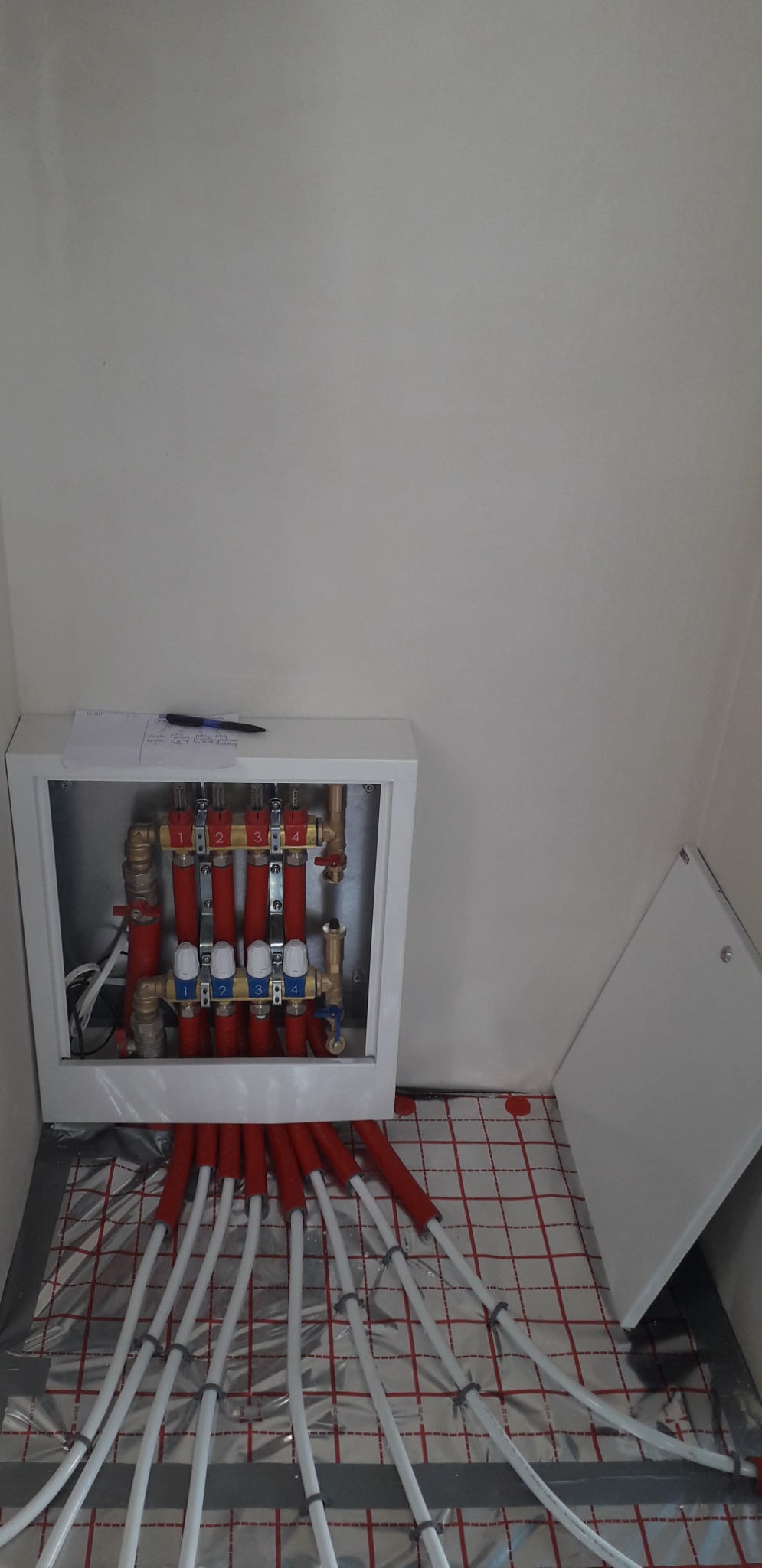 Rozdzielacz ogrzewania podłogowego w białej szafce, z widocznymi rurami na folii izolacyjnej z siatką. Notatka z długopisem na górze szafki. Ujęcie w rogu pomieszczenia.