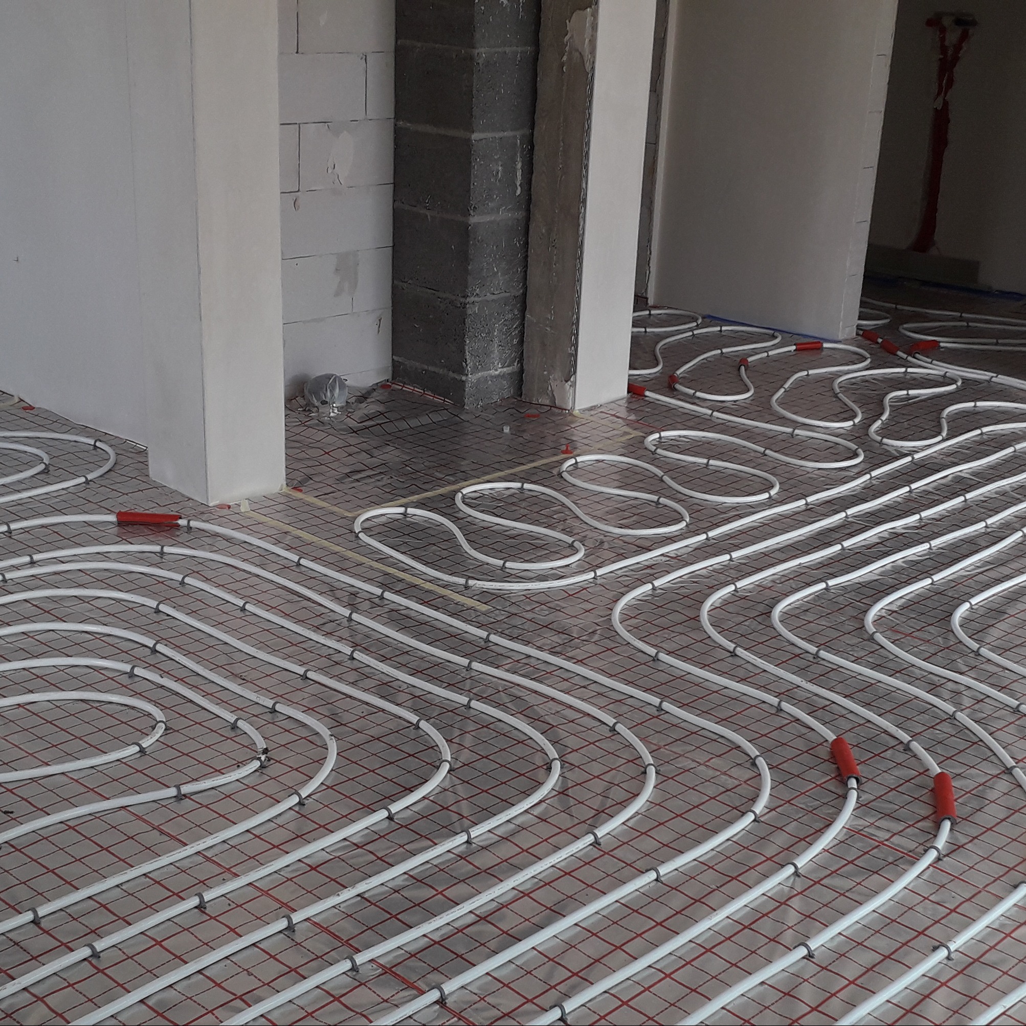 Białe rury ogrzewania podłogowego ułożone spiralnie na czerwonej siatce w surowym wnętrzu budynku z szarymi ścianami z bloków. Widok z góry, etap instalacji.