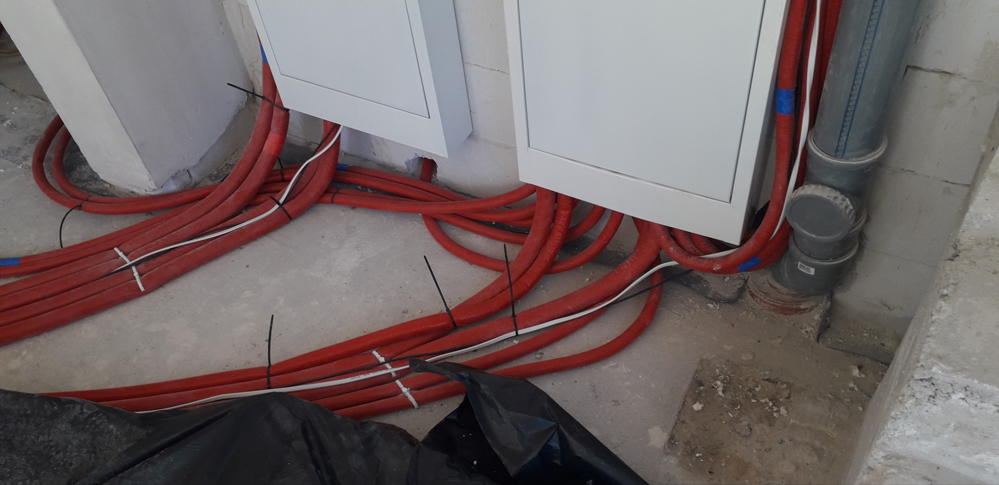 Czerwone rury instalacji grzewczej ułożone przy ścianie i szafkach w surowym wnętrzu. Widoczne fragmenty rur kanalizacyjnych i czarna folia zabezpieczająca.