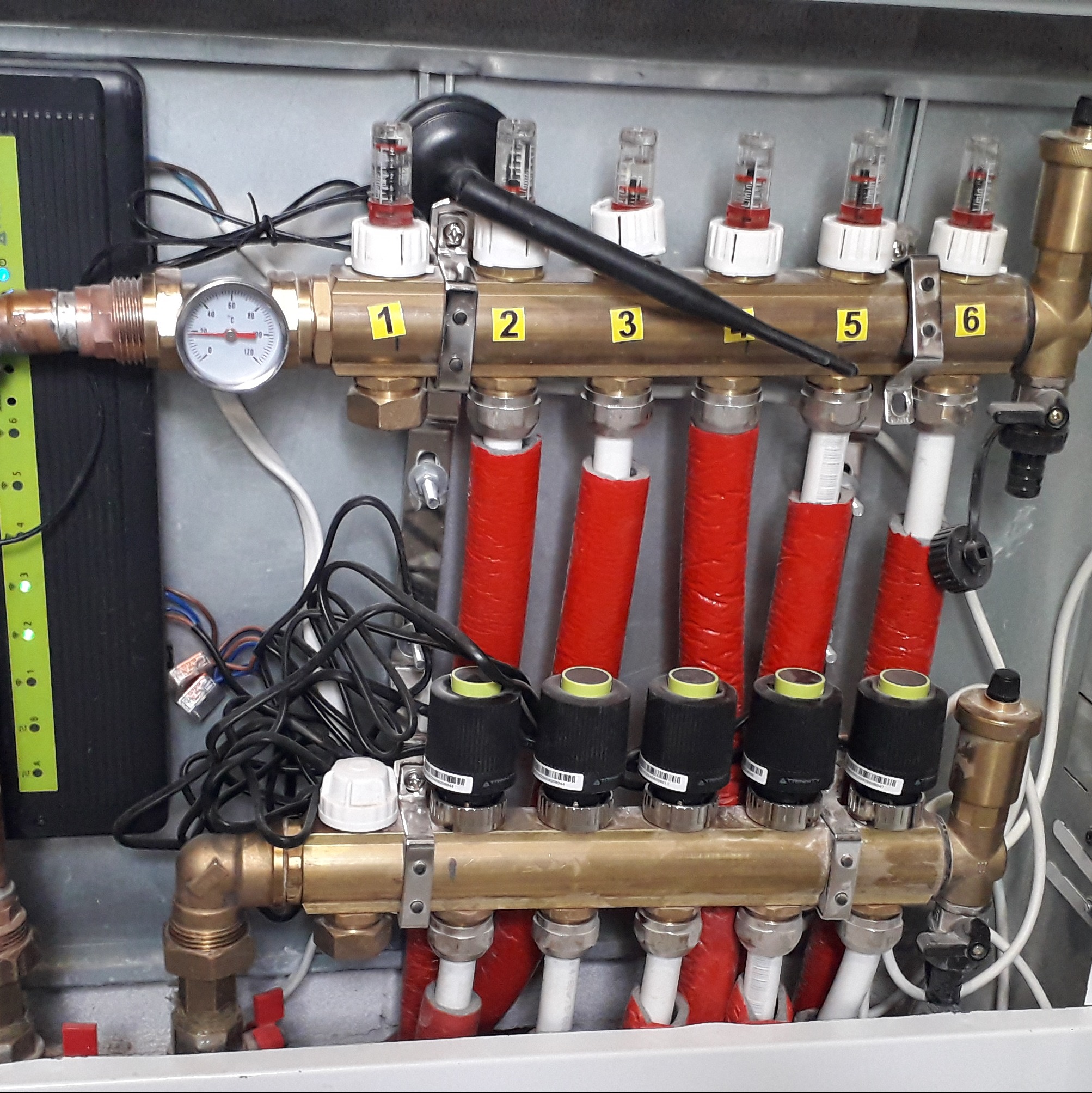 Rozdzielacz ogrzewania podłogowego w szafce, z numerowanymi sekcjami (1-6), czerwonymi rurami i czarnymi głowicami termostatycznymi. Widoczny manometr i przewody.