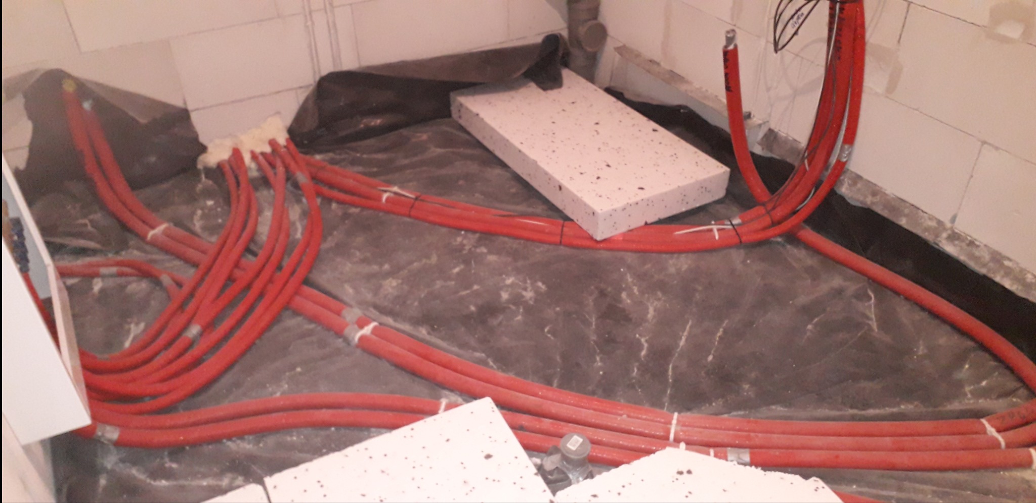 Czerwone rury ogrzewania podłogowego ułożone na czarnej folii izolacyjnej, gotowe do zalania posadzką. Widoczne fragmenty styropianu i ściany z bloczków.
