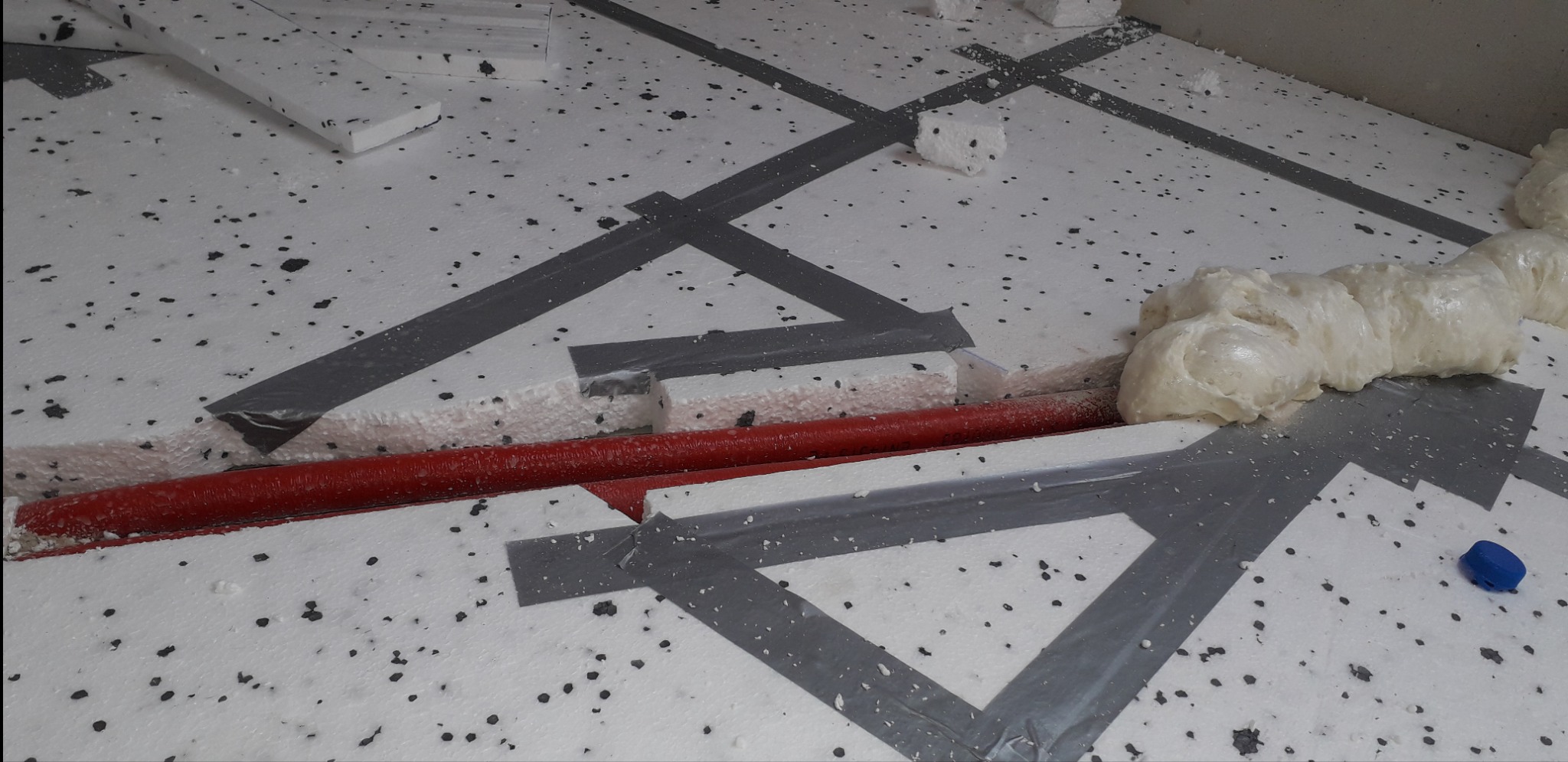 Czerwona rura ogrzewania podłogowego w izolacji styropianowej, zabezpieczona taśmą. Widoczna piana montażowa i fragment niebieskiego elementu.