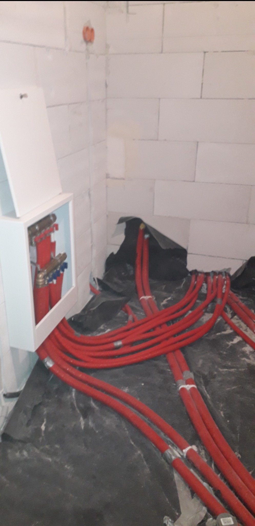 Rozłożone czerwone rury ogrzewania podłogowego na czarnej folii w surowym pomieszczeniu z białymi bloczkami, widoczna szafka rozdzielcza z zaworami.