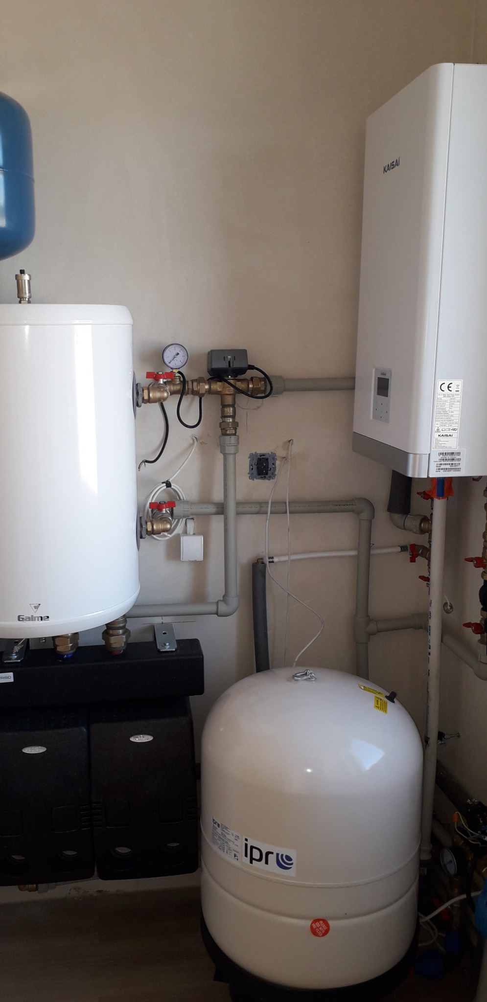 Kompletna instalacja grzewcza w kotłowni: biały zasobnik Galmet, piec gazowy Kaisai, naczynie przeponowe Ipr i orurowanie. Widoczny manometr i zawory.