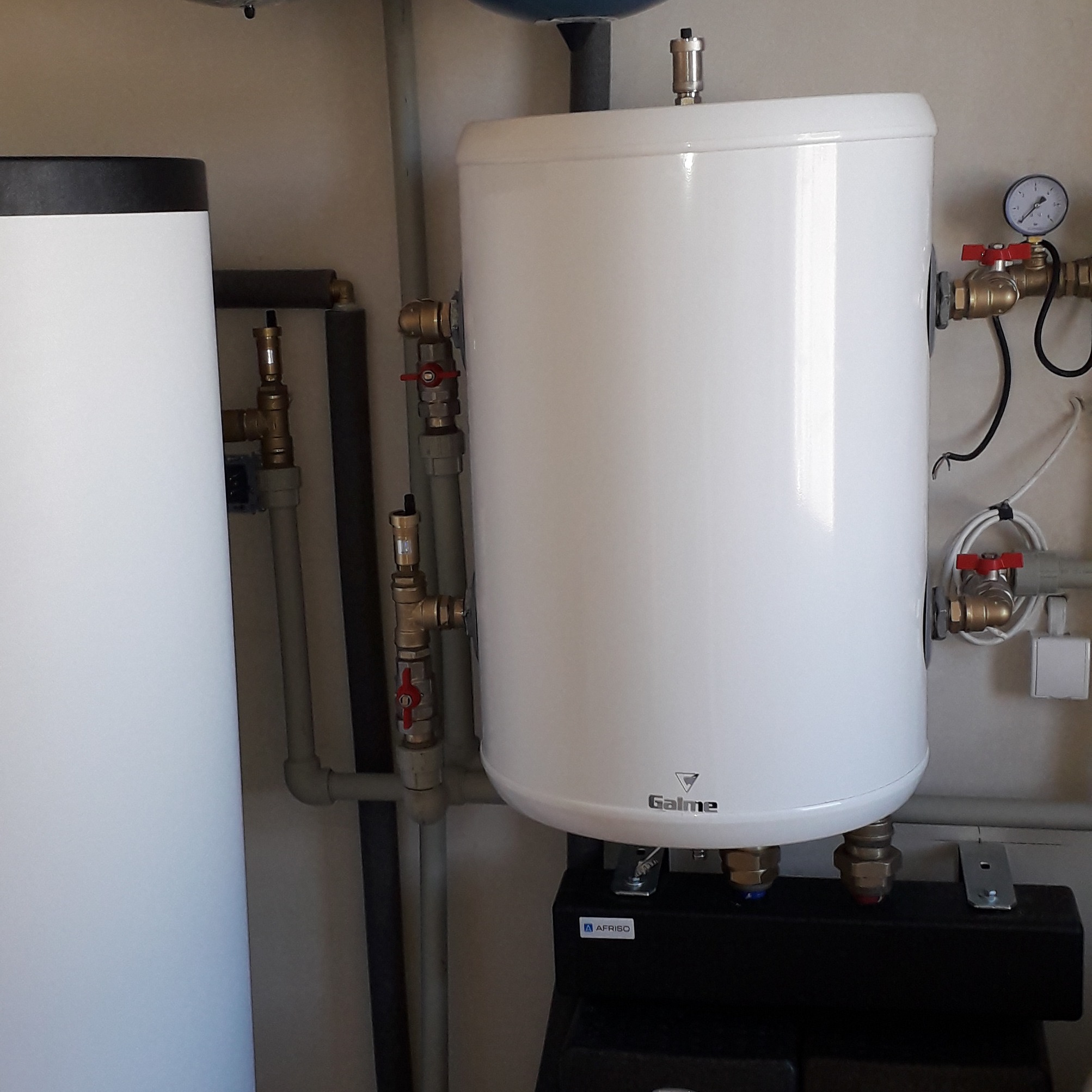 Biały, cylindryczny zasobnik ciepłej wody użytkowej marki Galmet, z widocznymi przyłączami hydraulicznymi i zaworami, zainstalowany w pomieszczeniu technicznym.