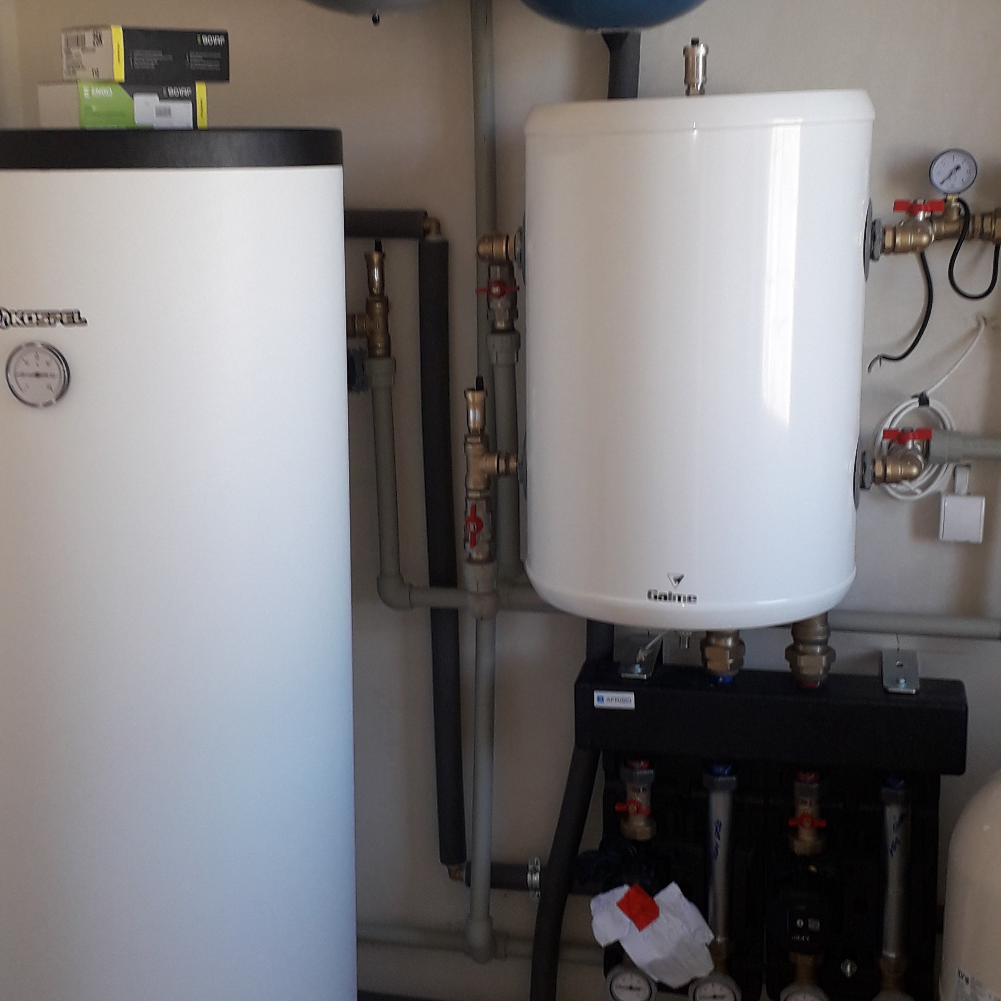 Biały bojler Galmet i zasobnik Kospel z oprzyrządowaniem: rury, zawory, manometry. Elementy instalacji grzewczej w kotłowni. Widoczne skrzynki i opakowania na górze bojlera.