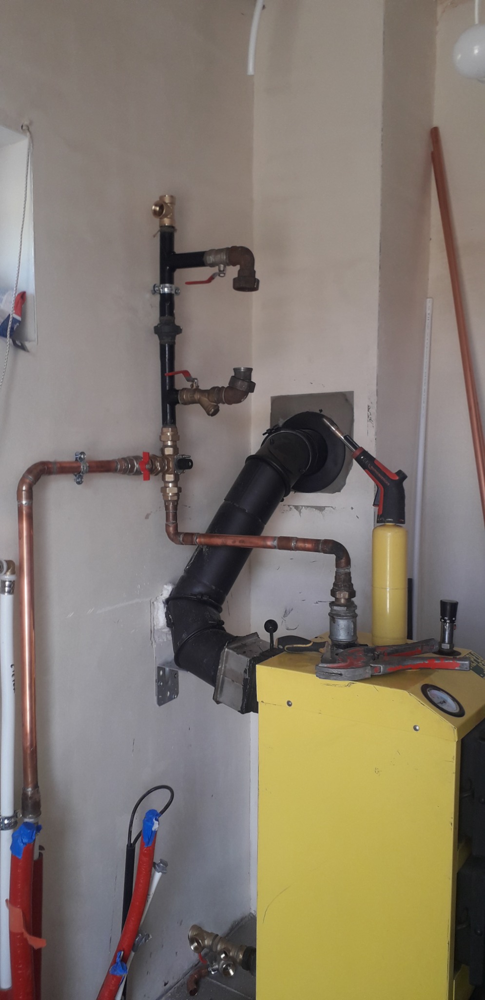 Żółty piec z podłączonymi rurami miedzianymi i czarnymi, zawory odcinające, narzędzia na piecu, instalacja wewnątrz budynku, widok z boku, oświetlenie dzienne.