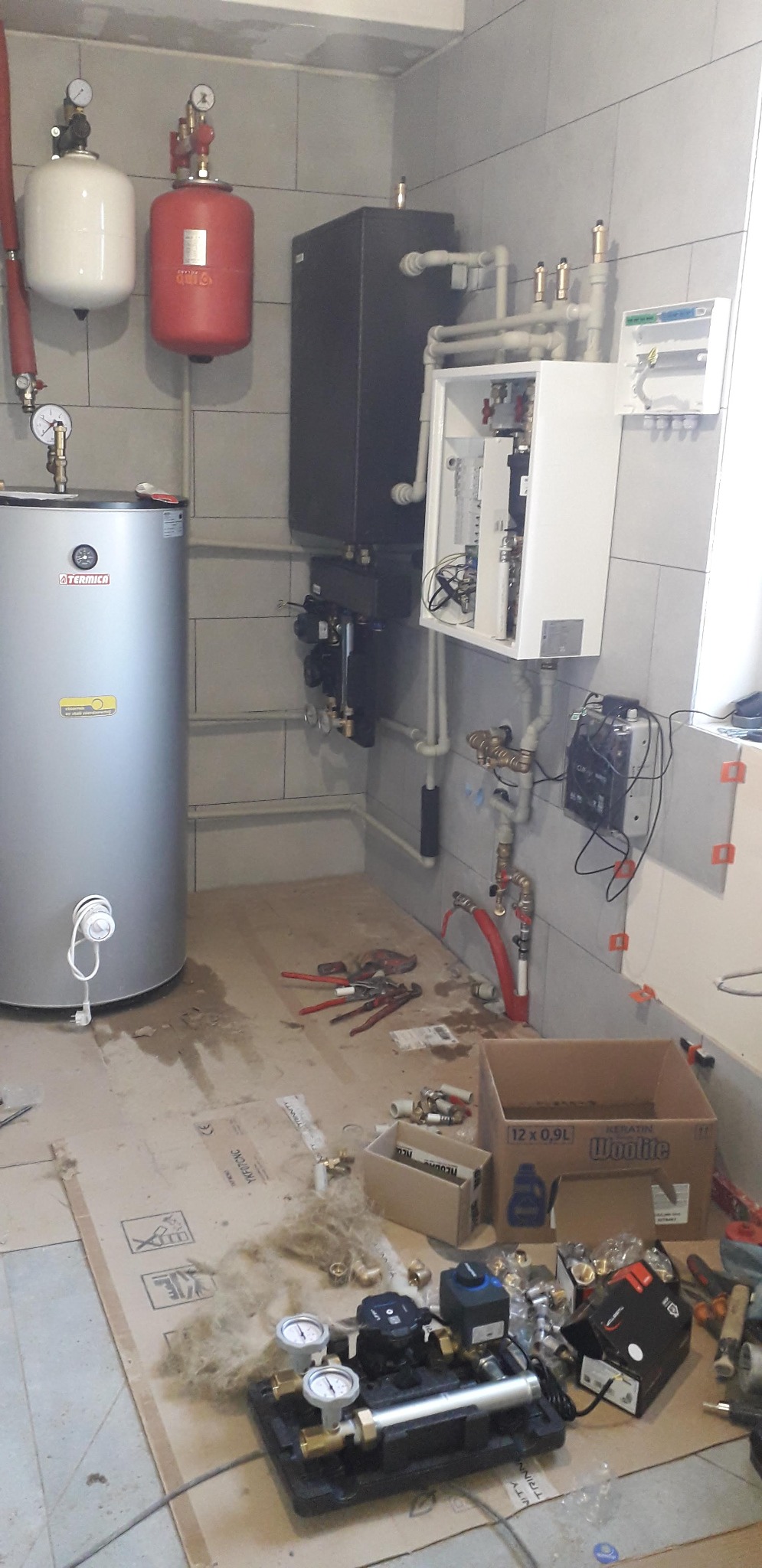 Wnętrze kotłowni w trakcie montażu: srebrny bojler Termica, czarny piec, biała skrzynka z instalacją, rury, manometry, narzędzia i kartony na podłodze.
