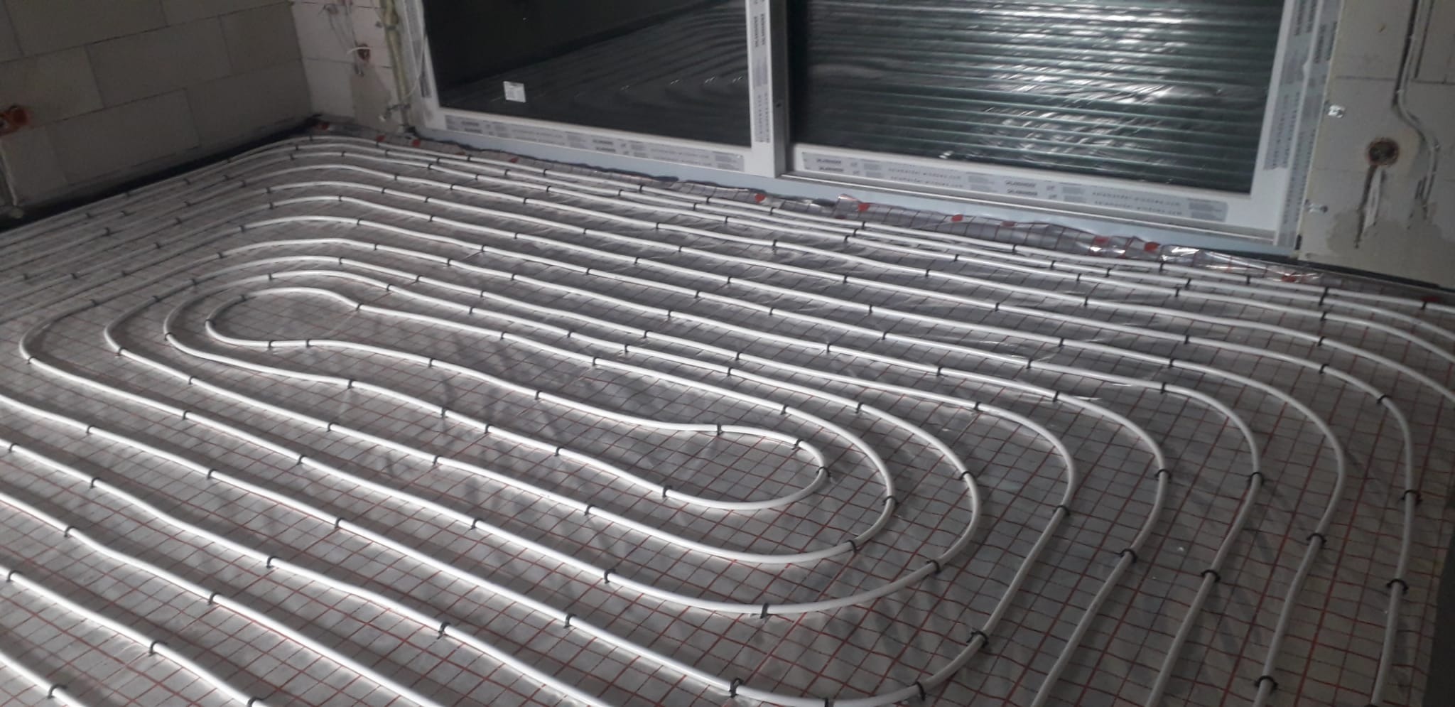 Białe rury ogrzewania podłogowego ułożone na folii z siatką w surowym wnętrzu, widoczne okno i fragment ściany z instalacją elektryczną.